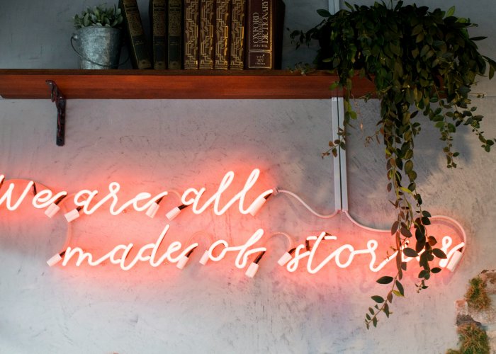 Leuchtreklame in Rosa mit der Aufschrift „We are all made of stories“ an einer Betonwand, darüber ein Holzregal mit alten Lexikabänden und Zimmerpflanze.