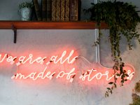 Leuchtreklame in Rosa mit der Aufschrift „We are all made of stories“ an einer Betonwand, darüber ein Holzregal mit alten Lexikabänden und Zimmerpflanze.