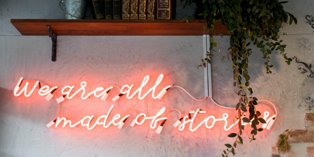 Leuchtreklame in Rosa mit der Aufschrift „We are all made of stories“ an einer Betonwand, darüber ein Holzregal mit alten Lexikabänden und Zimmerpflanze.